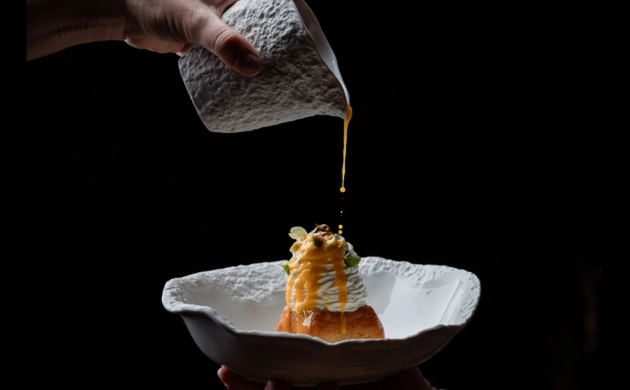 Dressage gastronomique d'un plat raffiné avec une main versant délicatement une sauce dorée sur une préparation dans une assiette texturée blanche, sur fond noir.