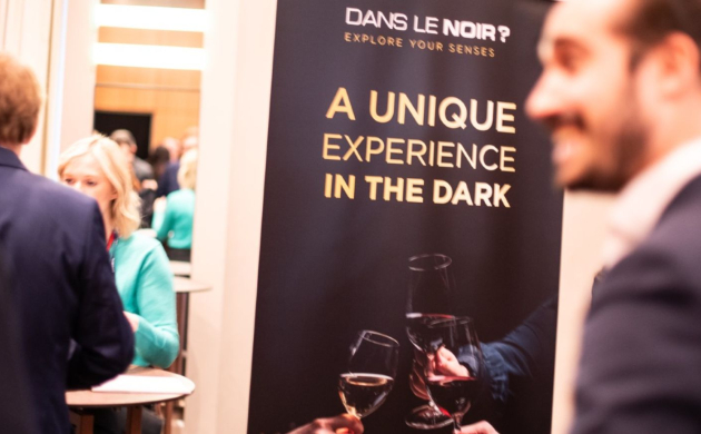 Des partenaires échangent lors d’un événement autour d’un panneau Dans le Noir ? affichant le slogan “A Unique Experience in the Dark”, verres en main.
