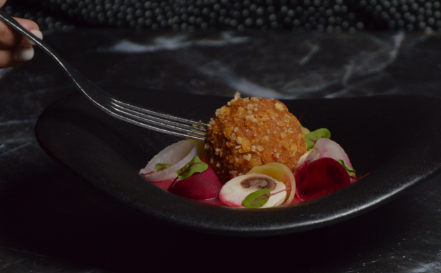 Assiette gastronomique dressée dans un bol noir avec croquette croustillante, légumes colorés et champignons, présentée sur table sombre